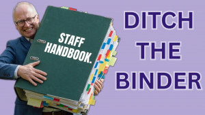 Smiling man in a suit holds a large green staff handbook binder with color-coded tabs on the side.